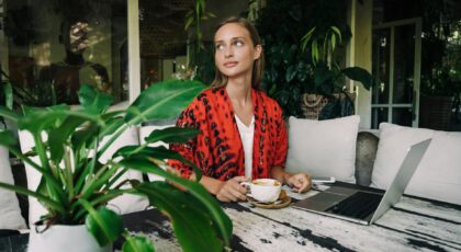 Woman sipping coffee while working on a laptop indoors with lush greenery.