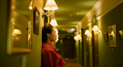 Profile of a woman in a red dress leaning against a hotel corridor under warm lighting.