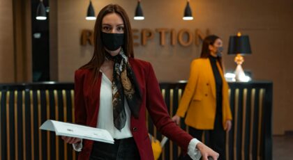 A woman in a mask stands at a hotel reception desk holding paper, welcoming service