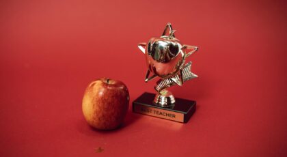 A 'Best Teacher' trophy alongside an apple on a vivid red background symbolizing education and recognition.