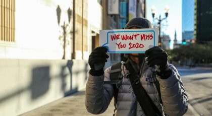 A person in winter clothing holds a sign reading 'We Won't Miss You 2020' on a city street.